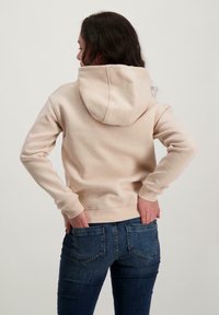Beige hoodie met een verstelbare capuchon, gemaakt van een zachte katoenmix, met ribgebreide manchetten en tailleband, gedragen met donkerblauwe jeans.