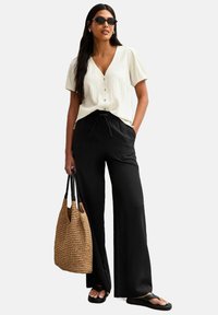 Blusa blanca texturizada con mangas cortas abullonadas, pantalones negros de pierna ancha con cinturilla, bolsa de mimbre marrón, chanclas negras y gafas de sol.