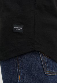 Černé tričko se zřetelnou námořnickou modrou nášivkou s nápisem „JACK & JONES EST. 1990“, v kombinaci s tmavými džíny s oranžovým prošíváním.