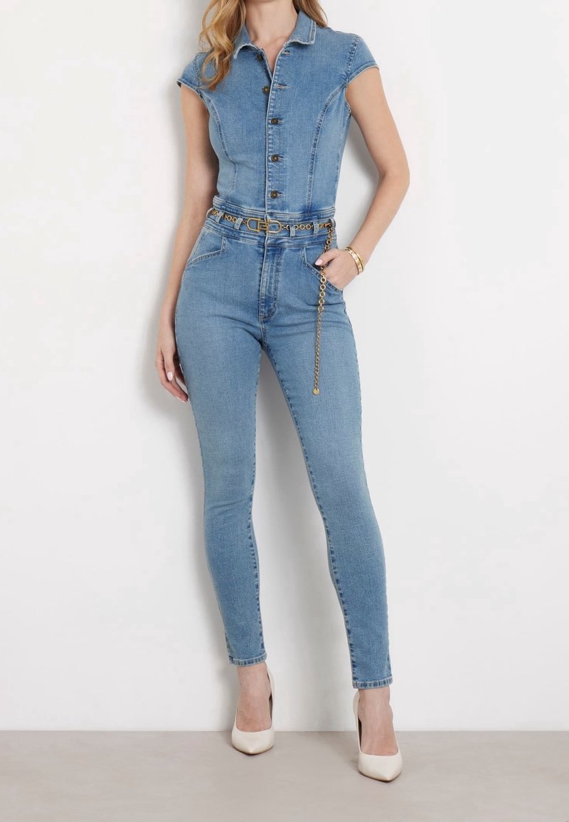 Femme portant une combinaison en denim bleu clair cintrée à manches courtes avec une ceinture chaîne dorée, associée à des chaussures blanches à talons hauts.