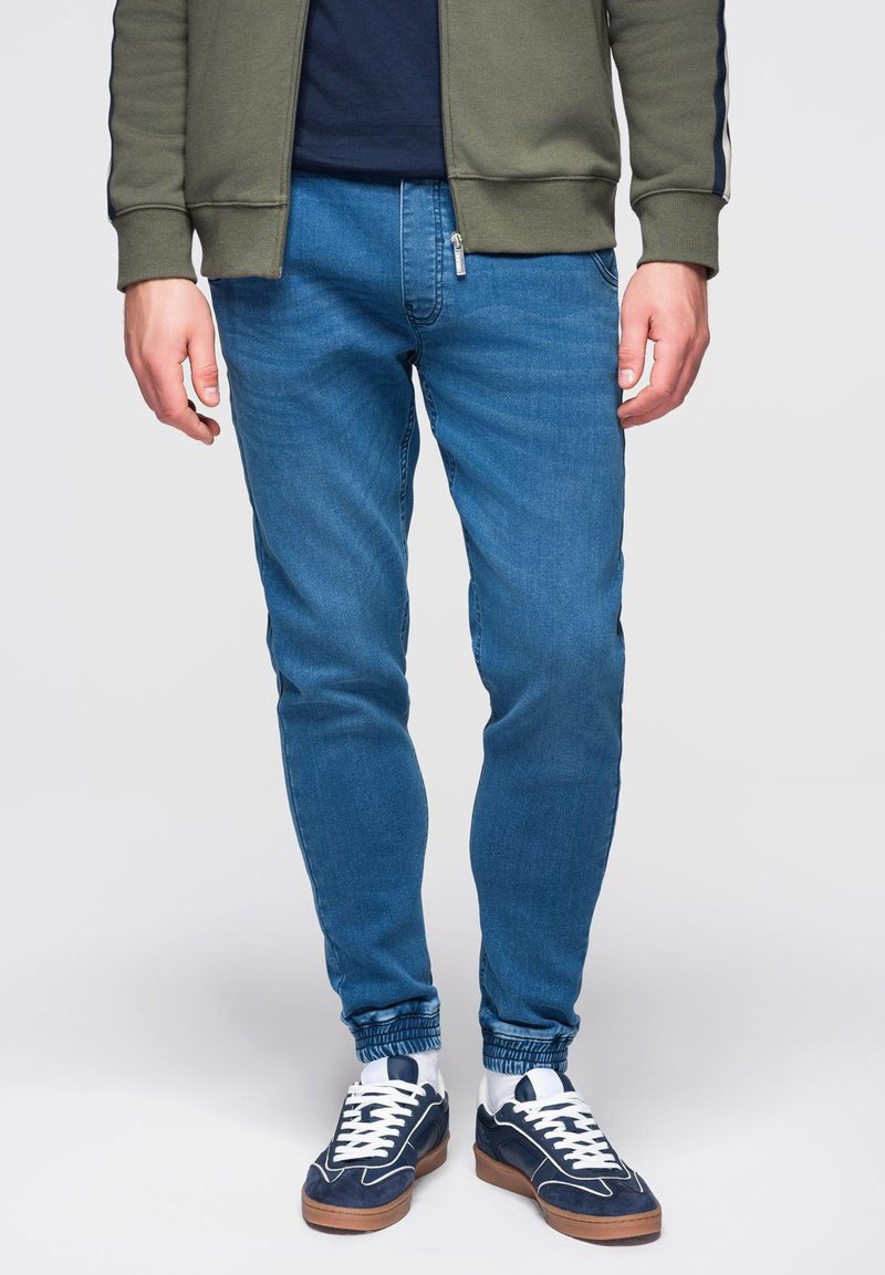 Jeans casual blu con vestibilità slim, caratterizzati da una tasca con zip, abbinati a sneakers bianche e blu navy e a un giubbotto bomber verde.