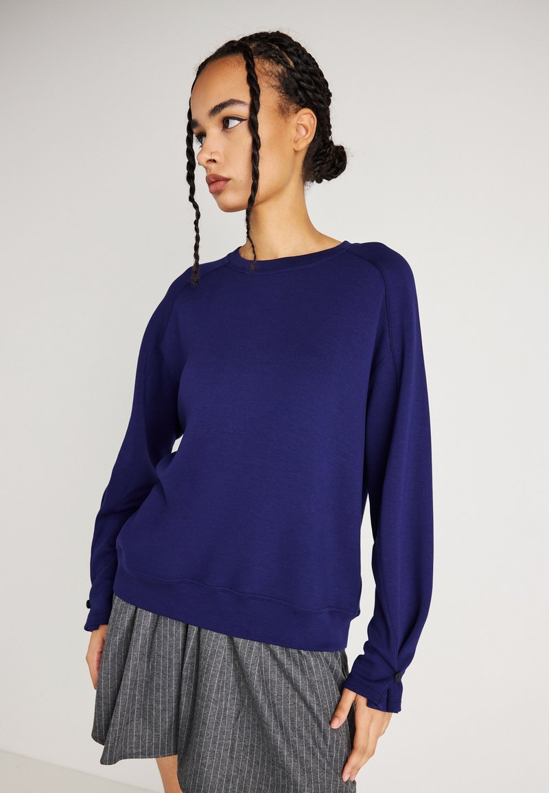 Tengerészkék pulóver hosszú raglán ujjakkal, kerek nyakkivágással és gombdíszítéssel az ujjak végén. Simára végzett pamut keverék anyag, laza szabás.