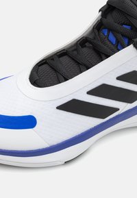 Κοντινό πλάνο ενός λευκού αθλητικού παπουτσιού με μαύρες κορδόνες, μαύρες διαγώνιες ρίγες, μπλε λεπτομέρειες και μια μπλε ρίγα κατά μήκος της σόλας.