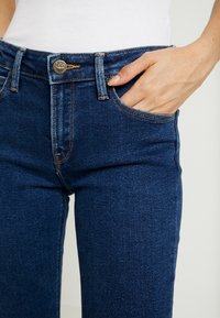 Mörkblå jeans i denim med hög midja, utrustade med metalldknapp, kopparfärgade sömmar och sidofickor. Tyget har en slät textur.