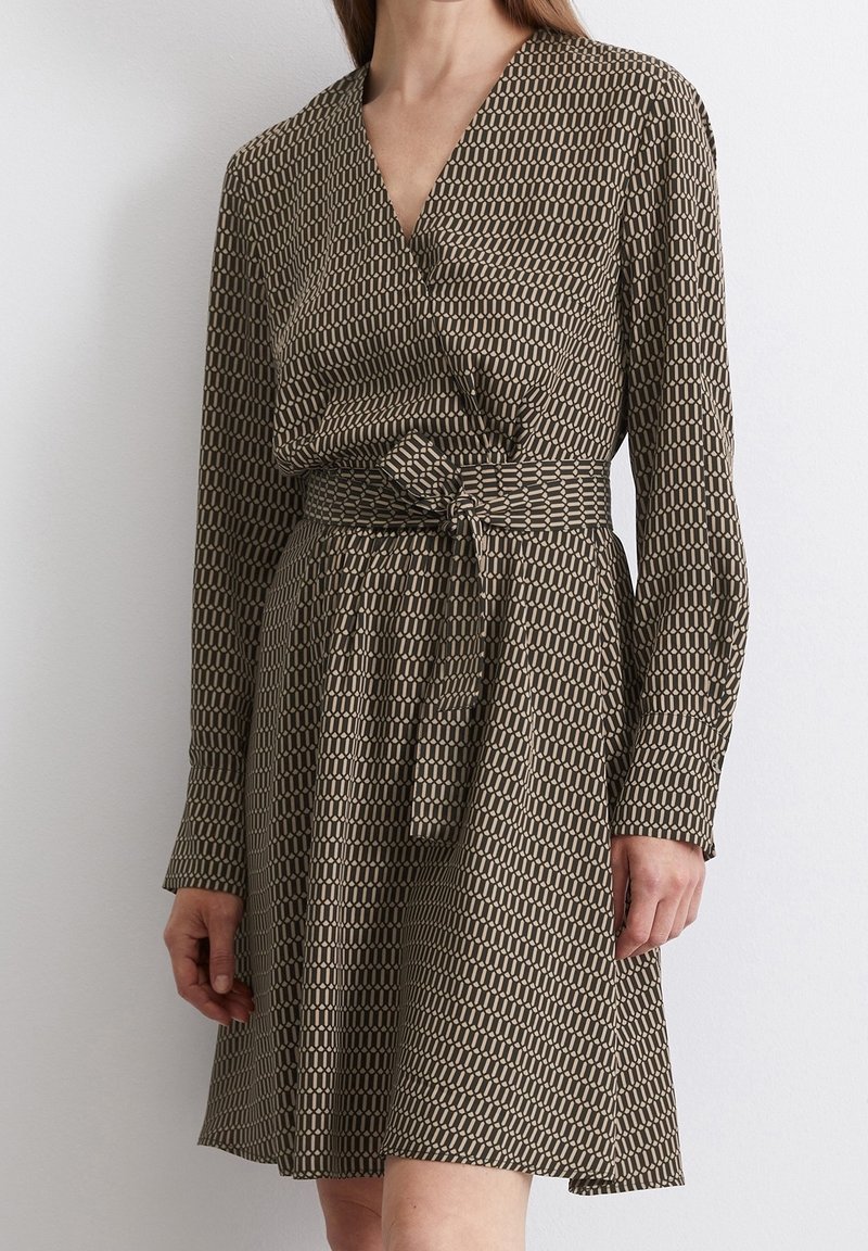 Femme portant une robe portefeuille à manches longues, longueur genoux, avec un motif géométrique et une ceinture en tissu assortie nouée à la taille.
