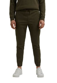 Homme portant un pantalon de jogging vert foncé avec des poignets élastiques et des baskets blanches Esprit, debout sur un fond blanc.