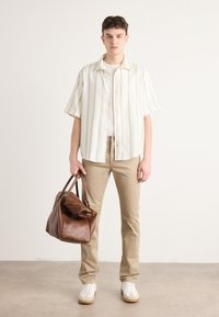 Model draagt een licht gestreept hemd met korte mouwen, beige broek en witte sneakers, en houdt een bruine leren weekendtas vast.