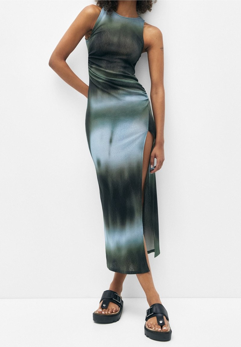 Femme portant une robe longue sans manches tie-dye avec une fente haute sur le côté et des sandales à plateforme noires avec de grandes boucles.