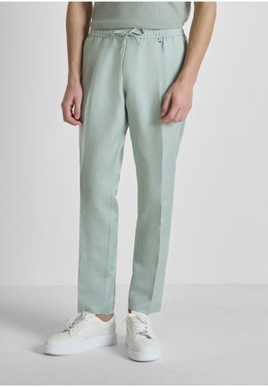 Hombre con pantalones de cordón verde claro y zapatillas blancas, de pie frente a un fondo liso gris claro.