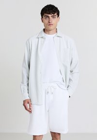 Giovane uomo che indossa pantaloni corti bianchi con coulisse, una maglietta bianca e una camicia a maniche lunghe a righe chiare sopra, su uno sfondo semplice e chiaro.