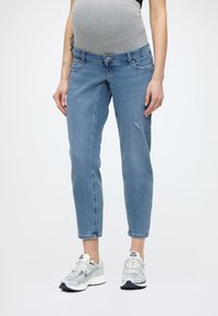 Εγκυμονούσες τζιν σε ανοιχτό μπλε τζιν ύφασμα, με στενή εφαρμογή, ελαφρώς φθαρμένο στυλ και ελαστική μέση. Συνδυάζονται με γκρι και λευκά αθλητικά παπούτσια.