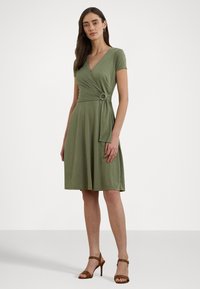 Vestido envolvente de manga curta, verde oliva, com um detalhe em nó na cintura, feito de tecido suave e combinado com sandálias de salto alto castanhas.
