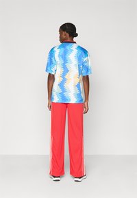 Mężczyzna z warkoczami stoi tyłem, ubrany w niebieską koszulę w wzory, czerwone spodnie szerokie z białymi paskami po bokach i sneakersy.