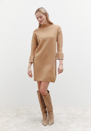 Robe pull beige tricotée avec un col montant et des manches évasées, présentant une texture subtile et des détails en perles au niveau des poignets.