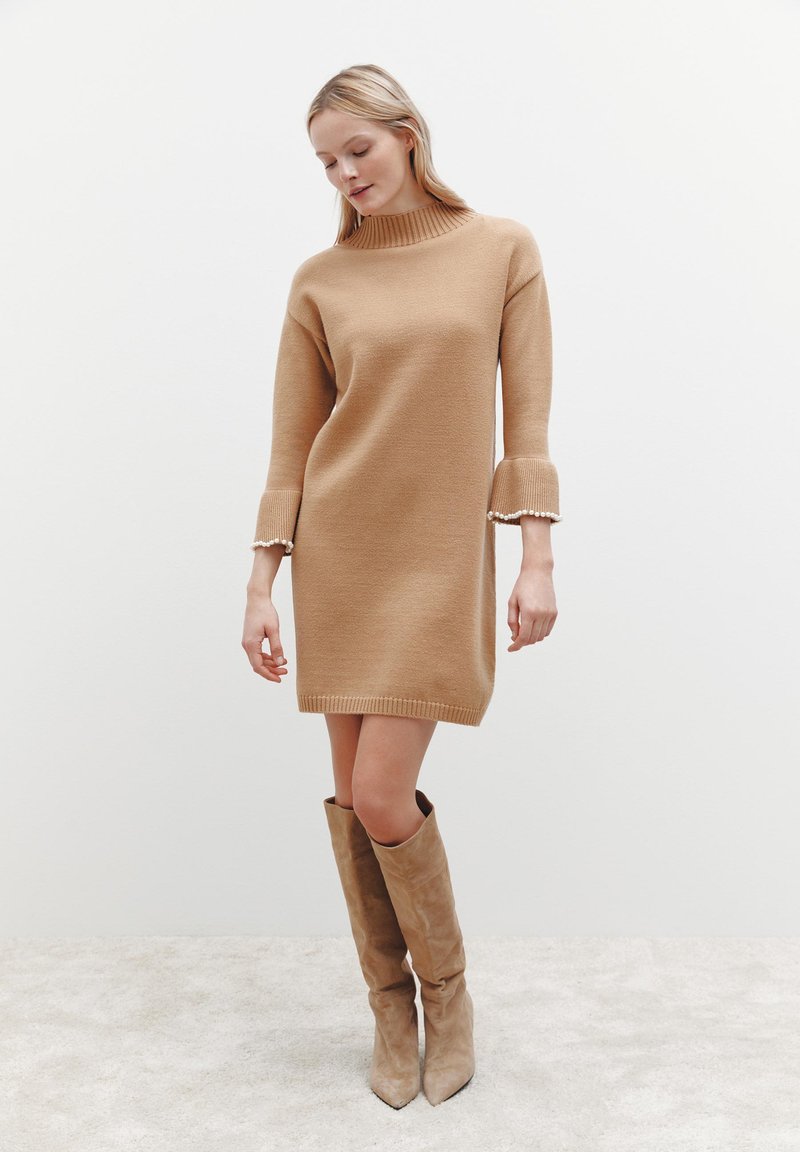 Beige gestricktes Kleid mit hohem Kragen und weit ausgestellten Ärmeln, das eine subtile Textur und Perlendetails an den Bündchen aufweist.
