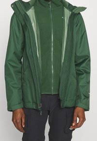 Giacca impermeabile verde con chiusura con zip, cappuccio e fodera in rete, caratterizzata da una texture uniforme e polsini regolabili. Abbinata a pantaloni neri.