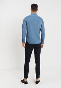 Jasnoniebieska koszula dżinsowa z zapięciem na guziki przy mankietach, zaokrąglonym dołem i kontrastowym przeszyciem, w połączeniu z czarnymi, dopasowanymi spodniami i czarnymi trampkami.