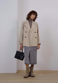 Chaqueta beige de doble botonadura, suéter de cuello alto gris, falda de mezclilla con abertura, botines marrón claro y un pequeño bolso negro.