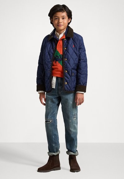 Marine quiltet jakke med brun kordfløjlskrage, orange sweater med grøn heste-design, slidte blå jeans og brune ruskindsankelstøvler.