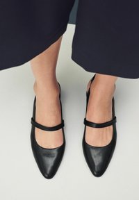 Las bailarinas de piel negra con tiras al tobillo presentan una punta puntiaguda, un escote cuadrado y una delgada correa en el tobillo para un ajuste seguro. Textura suave, diseño minimalista.