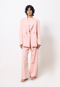 Rosa maßgeschneiderte Blazer mit doppelten goldenen Knöpfen, kombiniert mit passenden weit geschnittenen Hosen und hellrosa Sandalen. Glatte Stofftextur.