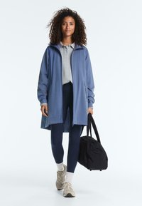 Femme aux cheveux bouclés portant un manteau bleu, un polo gris, des leggings bleu marine, des chaussettes blanches, des baskets et portant un sac de sport noir.