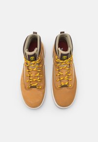 Bruna läderhög-top sportskor med gula snören, vadderad krage, gummisula och logopatch på tungan. Texturerad yta, stilren design.