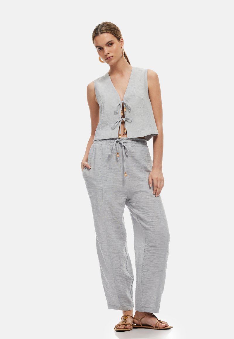 Femme portant un haut sans manches gris clair noué devant et un pantalon taille haute avec cordon assorti, avec des sandales marron sur fond blanc.