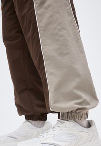 Prim-plan cu picioare purtând pantaloni jogger în culori maro și bej, cu mansete elastice, peste pantofi sport albi și șosete de culoare deschisă.