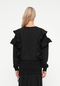 Hanorac negru cu accentuări mari din volane pe umeri și spate, cu un tiv ajustat și mâneci lungi. Textură netedă, design simplu.