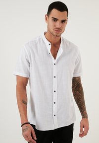 Cămașă albă cu mâneci scurte, realizată dintr-un material texturat, având un guler înalt și nasturi închiși la culoare pe partea din față. Design casual, potrivire lejeră.
