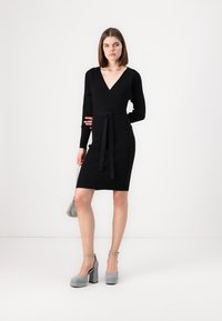 Vestido preto de malha canelada com decote em V profundo, mangas compridas e cintura atada. Combinado com saltos altos cinzas e uma bolsa prateada texturizada.