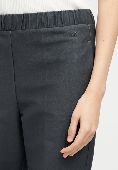 Κοντινή λήψη του χεριού ενός ατόμου που ακουμπά δίπλα σε σκούρο γκρι παντελόνι με ελαστική μέση και μικρό κεντημένο "MM" κοντά στην τσέπη.