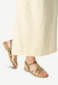 Gold and olive green strappy sandals with a flat sole, featuring adjustable ankle strap and wide crossover straps, set against a light fabric.