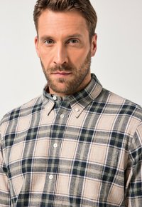 Koszula flanelowa zapinana na guziki, z beżowym, granatowym i zielonym wzorem w kratę. Materiał ma teksturowaną fakturę, z klasycznym kołnierzykiem i białymi guzikami.