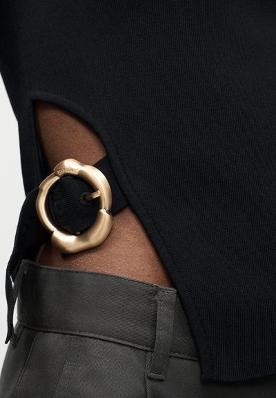 Μαύρο πλεκτό ένδυμα με πλευρική εγκοπή που στερεώνεται με μια υφασμένη χρυσή κυκλική αγκράφα, φορεμένο πάνω από σκούρο γκρι παντελόνι.