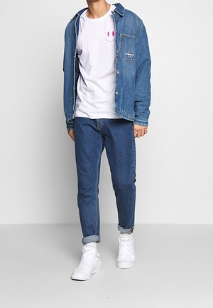 Denimjack met daaroverheen een wit T-shirt met roze afbeelding, gecombineerd met een blauwe spijkerbroek en witte sneakers. Casual straatstijl.