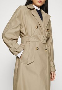 Cappotto trench beige con vita a cintura, design doppiopetto e bottoni grandi. Il tessuto ha una texture liscia e una silhouette strutturata.