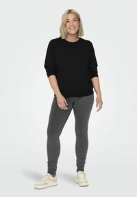 Maglione nero oversize con maniche lunghe, abbinato a jeans skinny grigi e sneakers bianche. Il modello è in piedi contro uno sfondo semplice.
