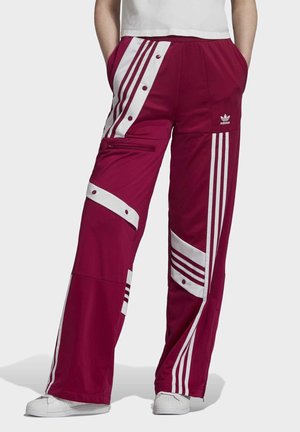 Pantalon de survêtement bordeaux Adidas avec des bandes blanches, boutons-pression, poche zippée et logo, porté avec des baskets blanches et un haut blanc.