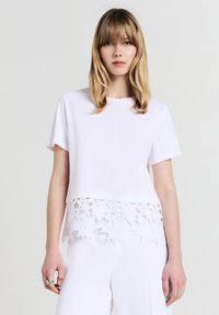 Donna con capelli biondi che indossa una maglietta bianca a maniche corte con orlo in pizzo floreale abbinata a pantaloni bianchi, su uno sfondo neutro.