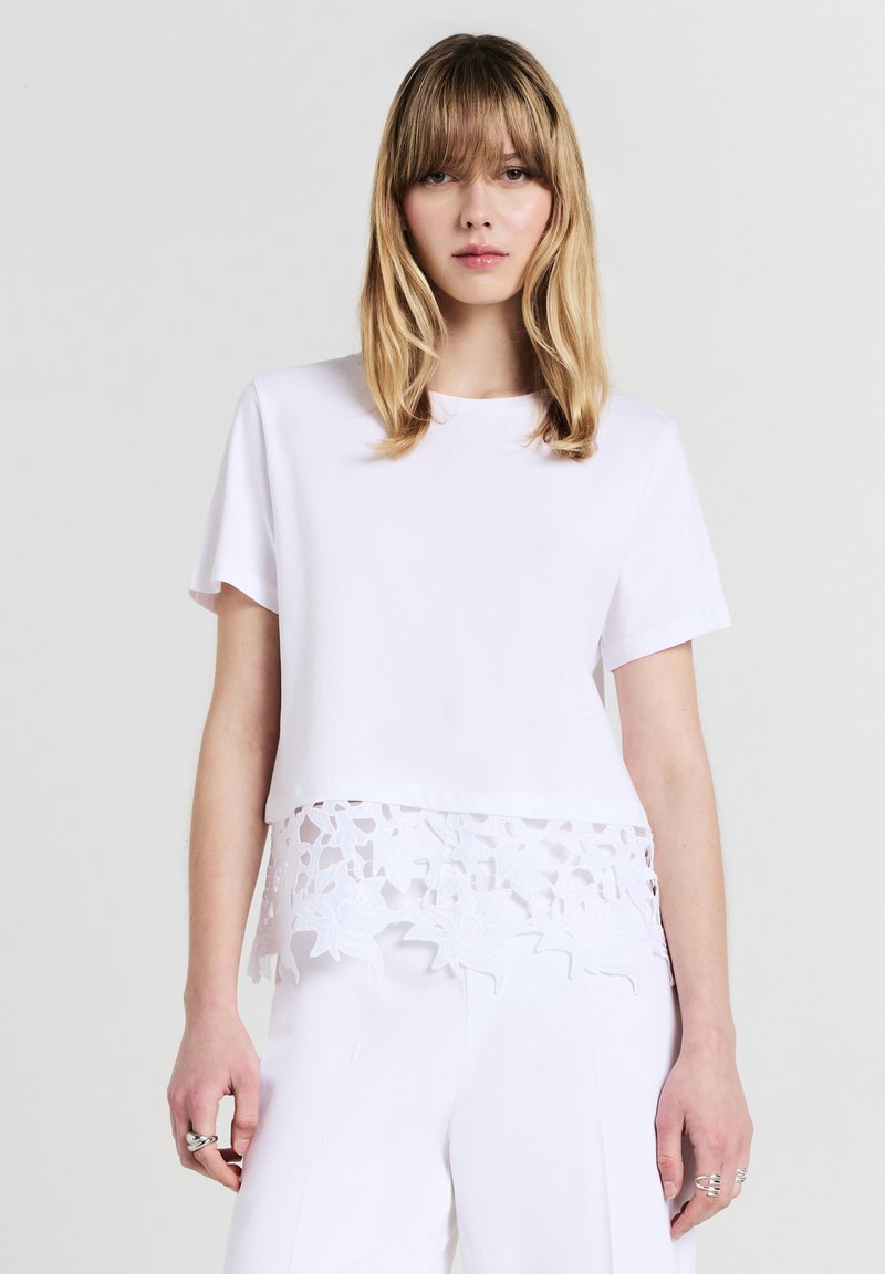 Donna con capelli biondi che indossa una maglietta bianca a maniche corte con orlo in pizzo floreale abbinata a pantaloni bianchi, su uno sfondo neutro.