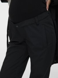 Tuvumā redzams cilvēks, kurš pielāgo vaļīgas melnas grūtniecības bikses virs melnas krekliņa, uzsverot komfortu un piegulšanu ap vēderu.