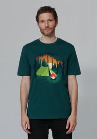 Donkergroene katoenen t-shirt met een ronde afbeelding van een tent, kampvuur en bomen tegen een oranje zonsondergang. 