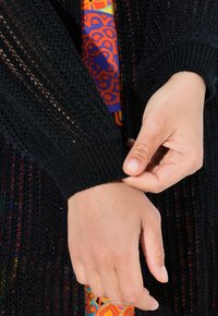 Černý pletený cardigan s otevřeným vzorem a manžetami s žebrováním; pod ním je viditelný barevný vzorovaný materiál. Ruce upravují manžetu.