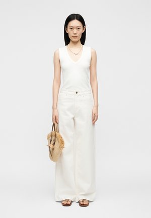 Γυναίκα που φοράει λευκή αμάνικη μπλούζα και λευκό παντελόνι με φαρδιά μπατζάκια, κρατώντας πλεκτή ψάθινη τσάντα με φούντες, στέκεται μπροστά σε απλό φόντο.