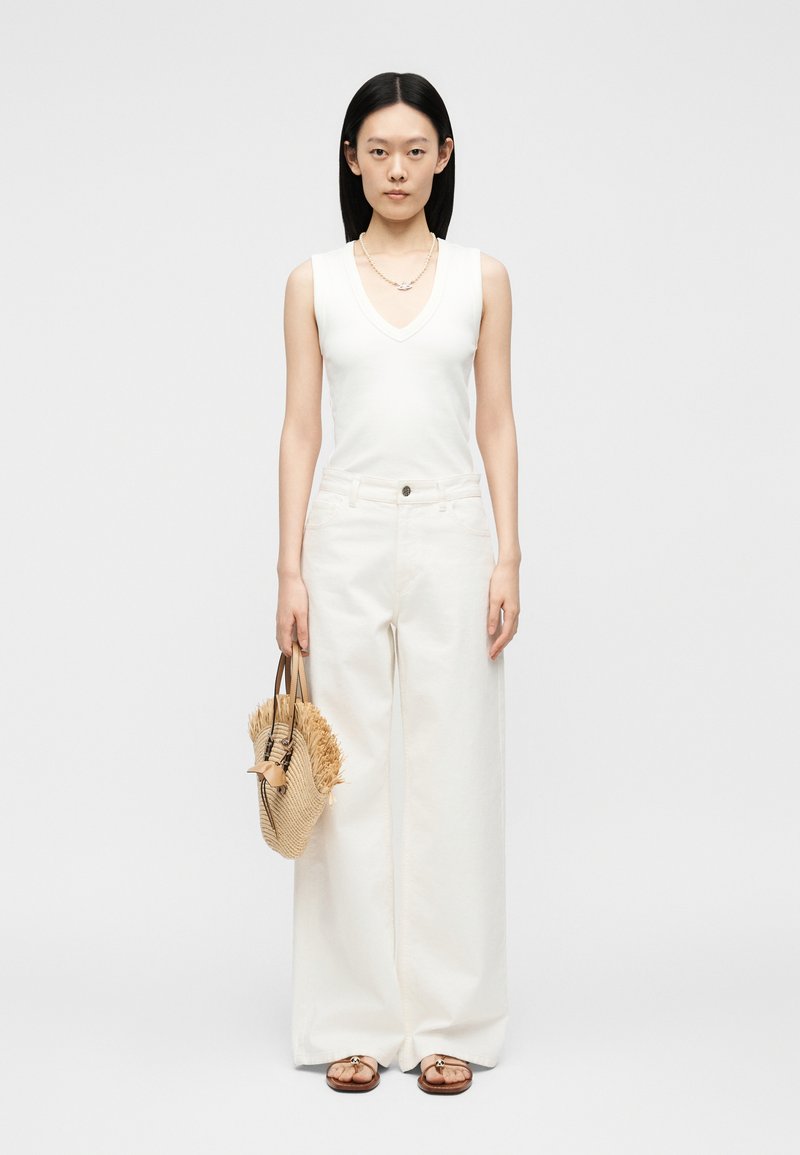 Γυναίκα που φοράει λευκή αμάνικη μπλούζα και λευκό παντελόνι με φαρδιά μπατζάκια, κρατώντας πλεκτή ψάθινη τσάντα με φούντες, στέκεται μπροστά σε απλό φόντο.