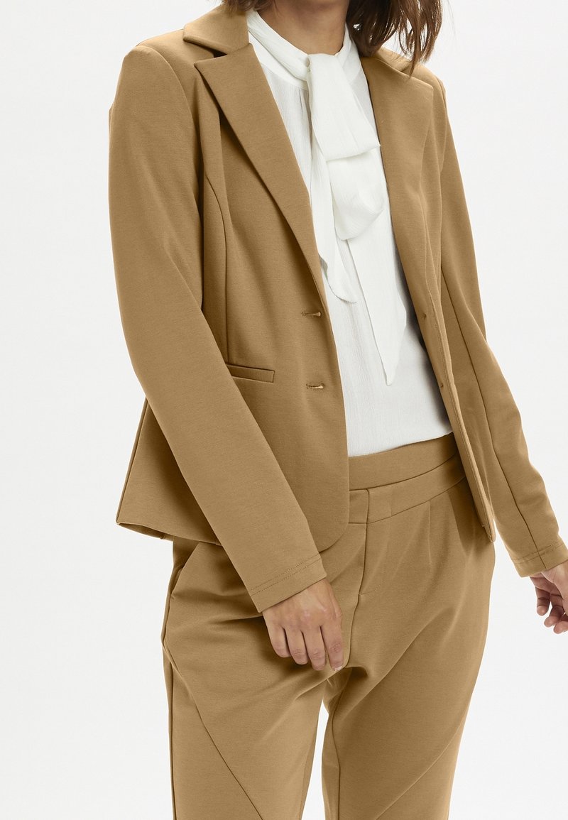 Femme portant un blazer et un pantalon couleur camel avec un chemisier blanc à nœud noué au col, debout sur un fond uni.
