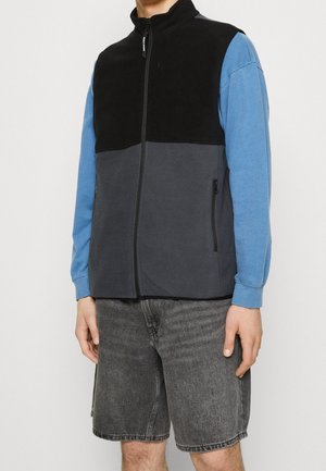 Homme portant un gilet polaire noir et gris zippé sur une chemise bleue à manches longues et un short en denim noir délavé, debout sur un fond blanc.