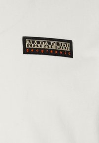 White cotton T-shirt featuring a black and orange rectangular embroidered label with "NAPAPIURI geographic" text. Smooth fabric texture.